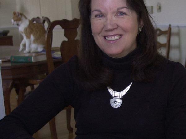 Joan Murray is smiling, and has black hair, at her back is a dining table with chairs with a white-brown cat, and a green book, she is wearing earrings, a choker locker, and a black turtle neck.