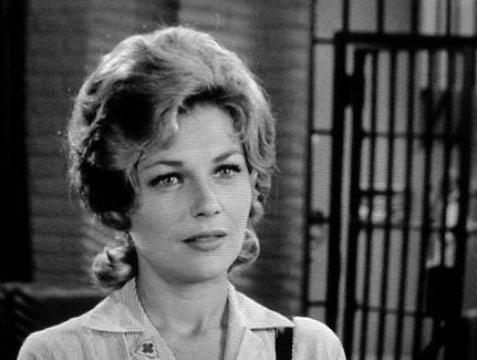 Joanna Moore in a scene from the 1960 tv series, The Andy Griffith Show. Joanna is smiling while looking at something, with wavy hair, a gate and a brick wall in the background. Joanna is wearing a blouse with a collar