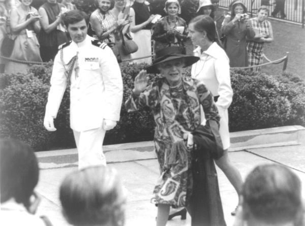 Alice Roosevelt Longworth wearing a dress and a hat with her granddaughter Joanna Sturm wearing a white dress escorted by the White House Social Aide at the crowd.