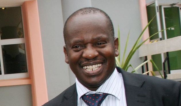 Job Ndugai with a big smile while wearing a black coat, striped long sleeves, and blue polka dot necktie