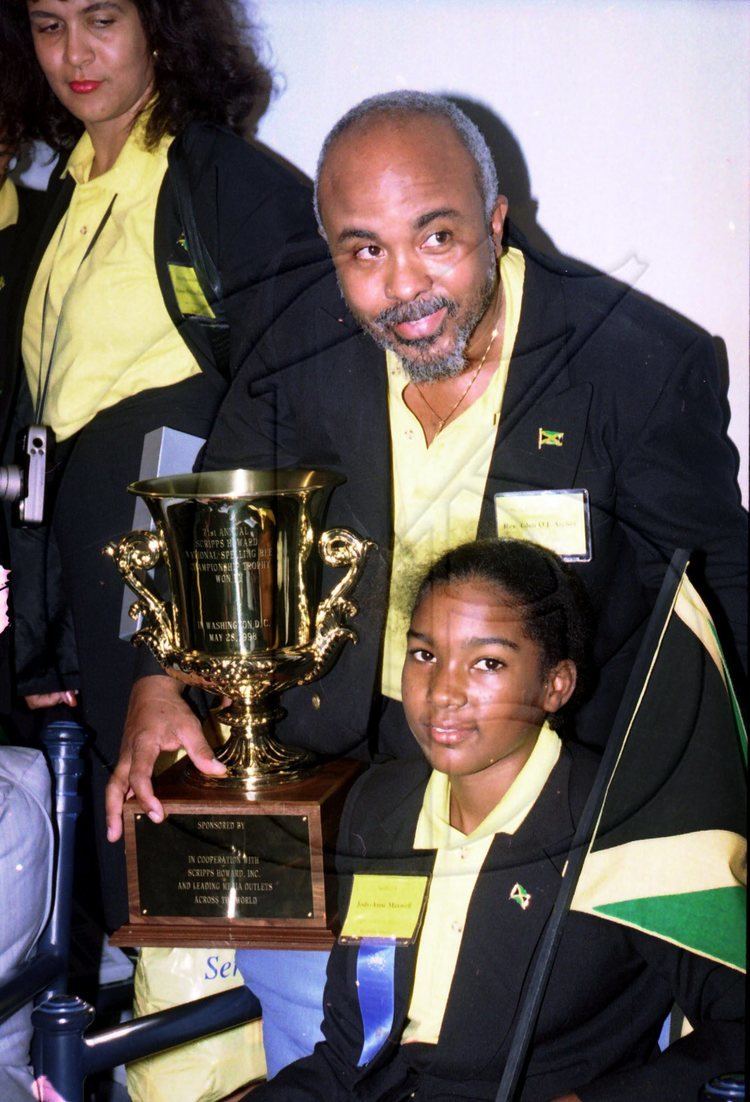 Jody-Anne Maxwell smiling, wearing a yellow shirt and a black jacket with a big trophy beside her.