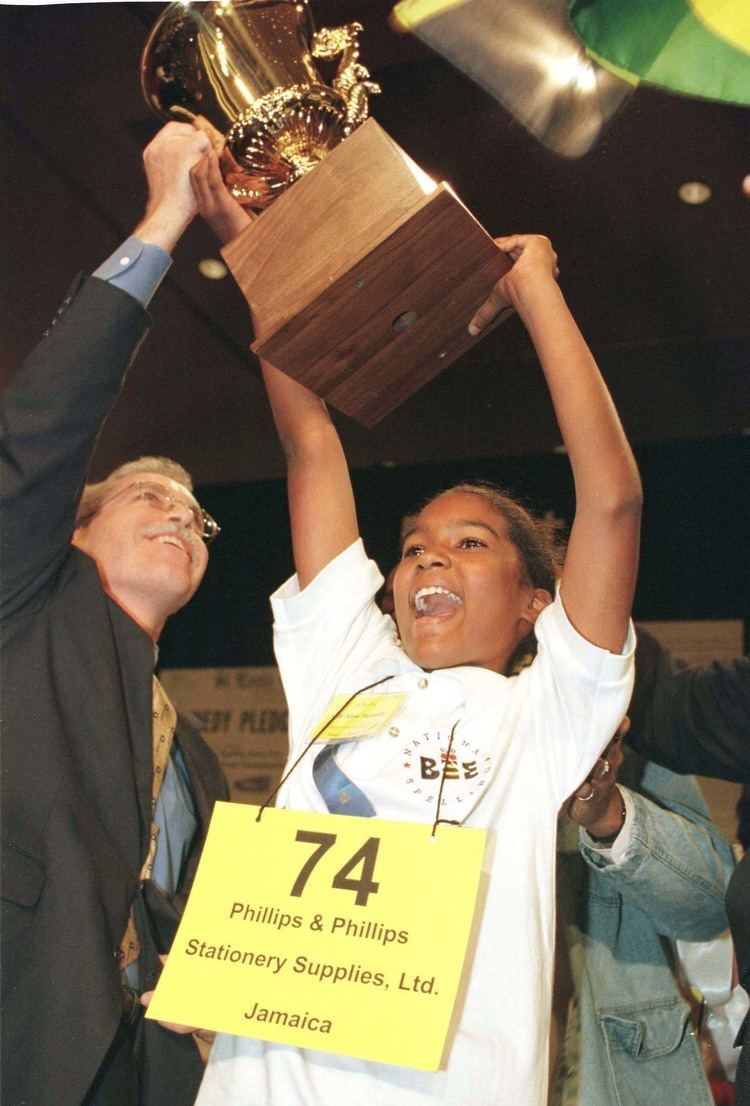Jody-Anne Maxwell wearing a white polo shirt while holding a big trophy for winning the 1998 Scripps National Spelling Bee.