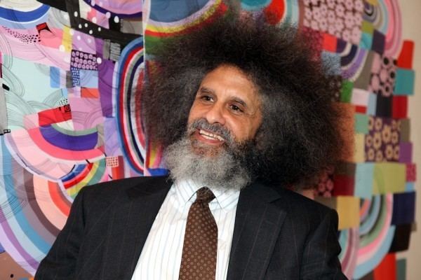 Jody Armour smiling and wearing a black coat over a white suit and brown tie with his trademark thick hair and beard.
