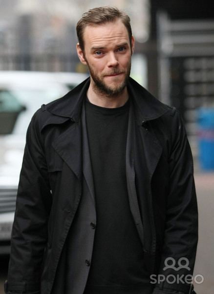 Joe Absolom biting his lips with mustache and beard while wearing a black coat, black vest, and black t-shirt