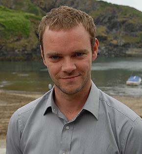 Joe Absolom with a tight-lipped smile while at the lake and wearing a gray long sleeves