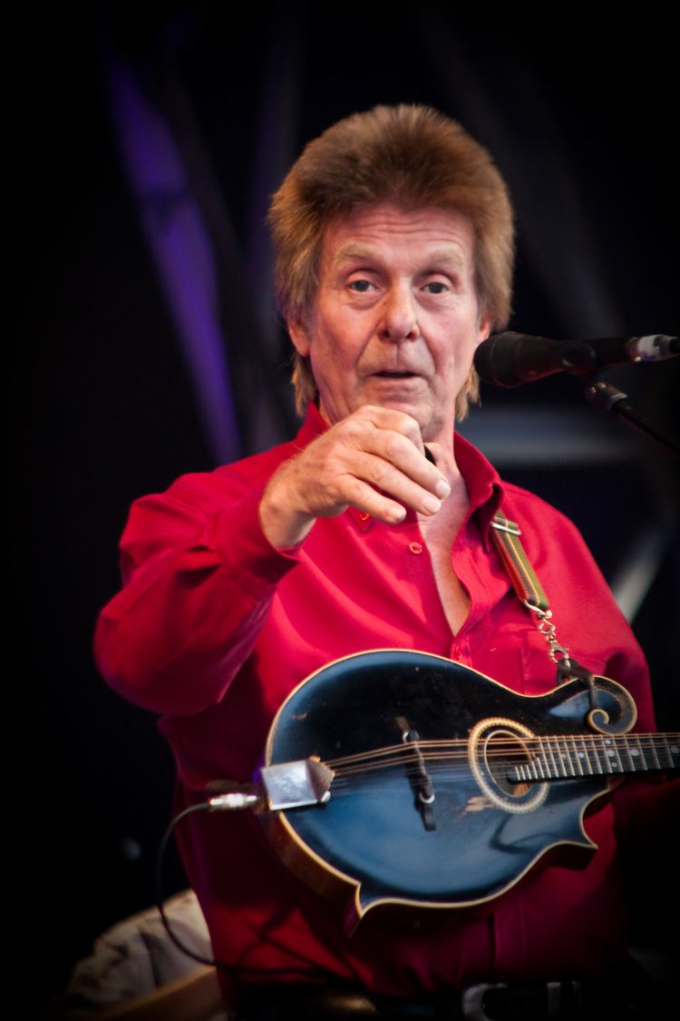 Joe Brown with blonde hair, wearing red long sleeves and carrying a guitar.