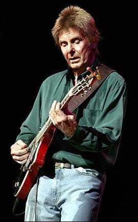Joe Brown with blonde hair, wearing green long sleeves and blue jeans while playing guitar.