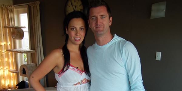 Joe Cotton and her husband Daniel Shields are smiling. Joe with long black hair and wearing a white spaghetti dress while her husband wearing a light blue long sleeve shirt.
