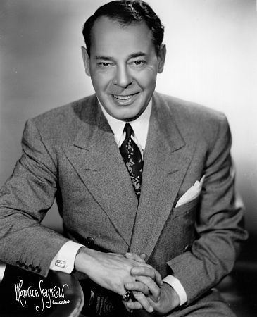 Joe E. Lewis smiling while wearing a coat, long sleeve, and necktie