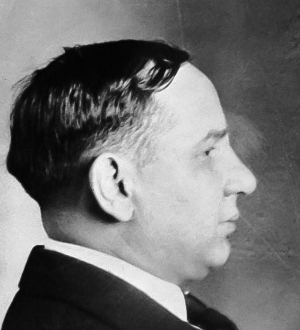 A portrait of Joe Masseria posing side view and wearing a black suit over a white polo shirt and gray tie.