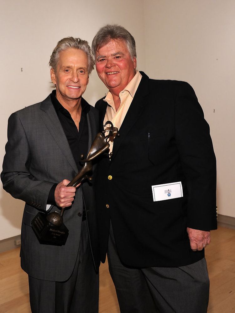 Michael Douglas wearing gray coat and black long sleeves while happily holding a trophy and Joel Douglas wearing black coat and yellow long sleeves