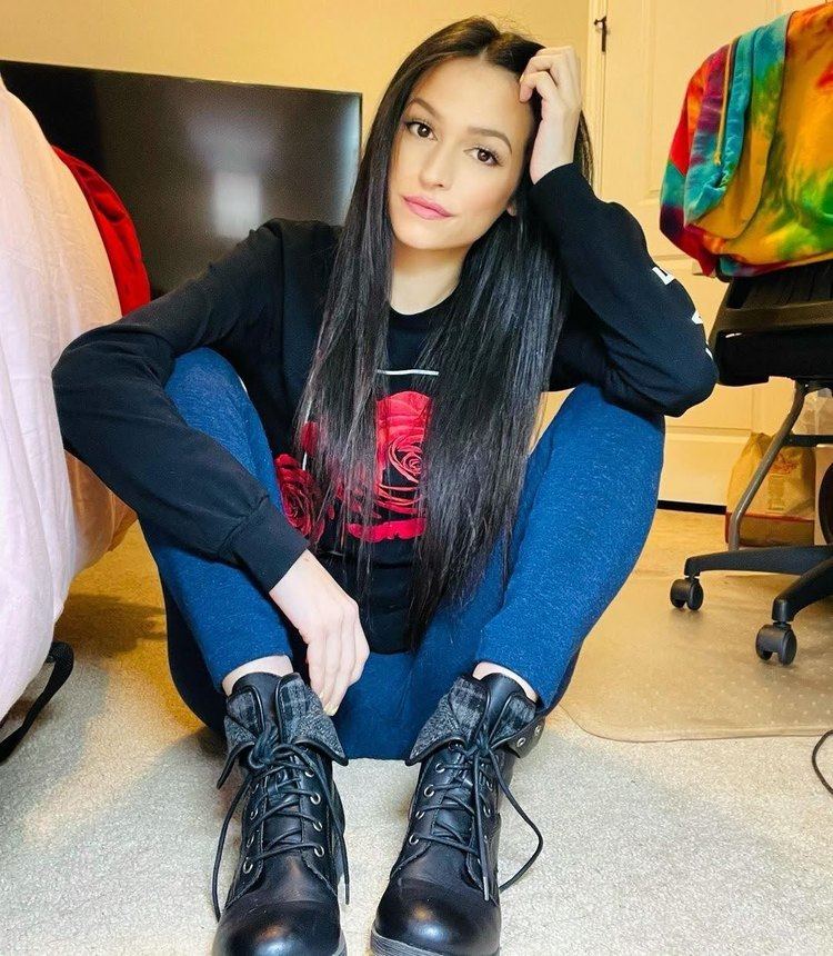 Joelle Grieco sitting on the floor while hand on her head and wearing a black sweater with a red rose print, denim pants, and black shoes