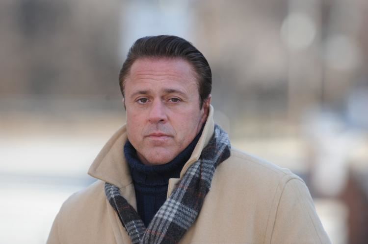 John Alite wearing a brown coat, a blue shirt with a scarf on his neck.