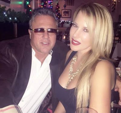 John Alite wearing a suit, a white shirt, and eyeglasses, and beside him a lady wearing a sexy black dress with long blonde hair and wearing a necklace.