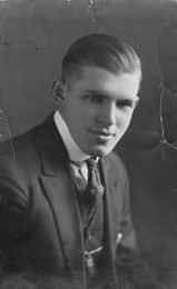 John Augustus Larson with a tight-lipped smile, wearing a black coat over white long sleeves, and a black necktie.
