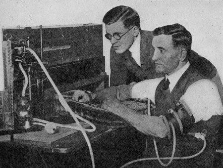 John Augustus Larson with a man looking at the polygraph, wearing eyeglasses, coat, and white shirt.