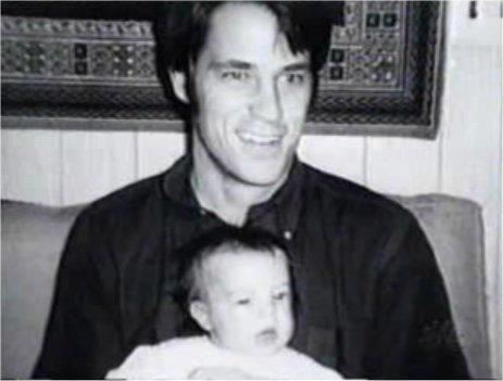 In black and white, has a room with white wooden walls with a frame hanging, John Bennett Perry is smiling, sitting on a white sofa, holding a Mathew Perry (baby) wearing white shirt in front, he has black hair and is wearing a black polo shirt.