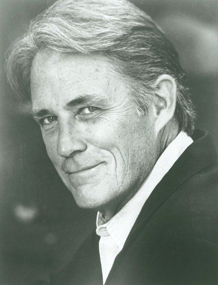 In a black and white John Bennett Perry is smiling, has dark-white hair wearing a white polo under a black coat.