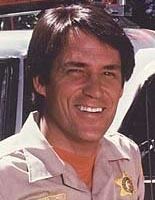 John Bennett Perry is smiling, standing in front of a white car, has black hair wearing a gray polo shirt with a yellow star printed on his left chest.