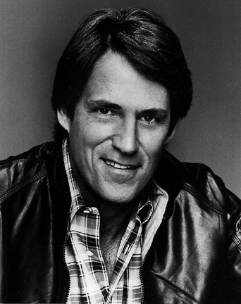 In black and white, John Bennett Perry is smiling, has black hair wearing a checkered polo under a black leather jacket.