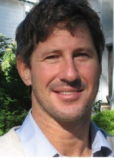 John Bowe smiling with messy brown hair and bushes in the background, a well-reputed journalist. He is wearing a white jacket over a light-blue collared shirt