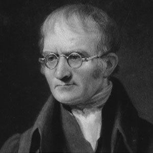 A portrait of John Dalton looking serious, with eyeglasses wearing a scarf under a black coat