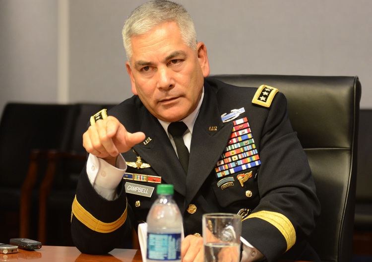 John F. Campbell sitting on the chair while pointing to someone and wearing a military coat with badges, white long sleeves, and necktie