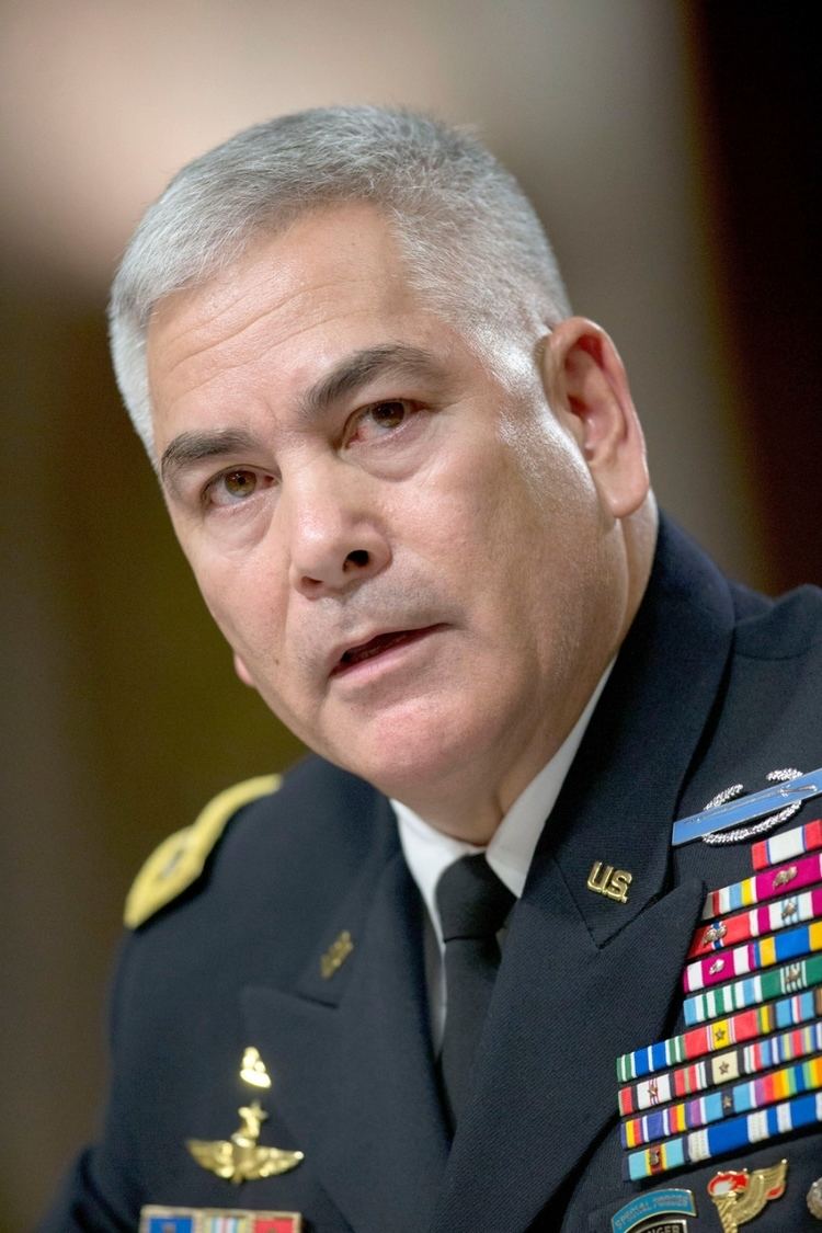 John F. Campbell looking afar while wearing a military coat with badges, white long sleeves, and a black necktie