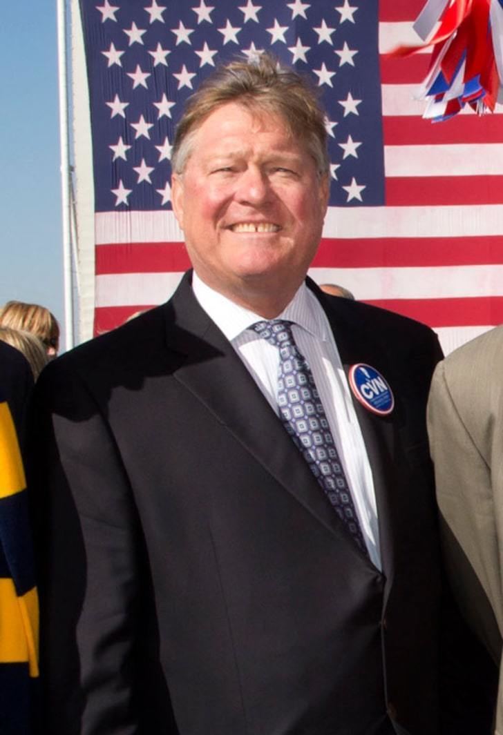 John Gardner Ford, smiling with a brown and white hair, with the American flag in his background, wearing a white long sleeve polo under a black coat, a dark purple and white necktie with a blue badge on his left chest with the "CVN" word written on it