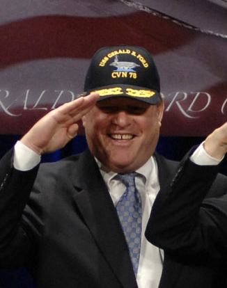 John Gardner Ford, smiling with a hand salute gesture, with a black cap with yellow printed words like "CVN 78" on his head and wearing a white long sleeve polo under a black coat, a dark purple and white necktie