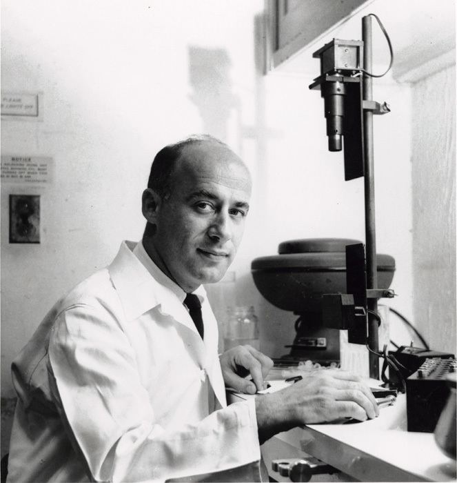 John Gofman with a tight-lipped smile beside laboratory apparatus at Donner Lab, Berkeley, California. John is wearing a white laboratory gown, white long sleeves, and a black necktie.