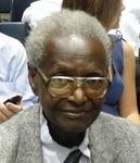 Dr. John Albert Musselman Karefa-Smart is smiling and has white curly hair, wearing eyeglasses, white long sleeves, and a black necktie under a gray suit.