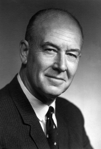 John Tuzo Wilson smiling while looking at something in a grayish background, bald, and wearing a shirt and a printed necktie underneath his coat.