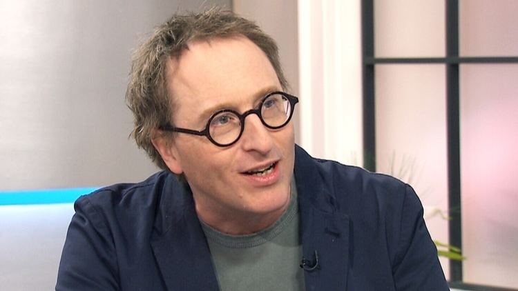 Jon Ronson is serious, has black hair, mouth half open, and wears black eyeglasses, and a gray shirt under a dark blue suit with a mic.