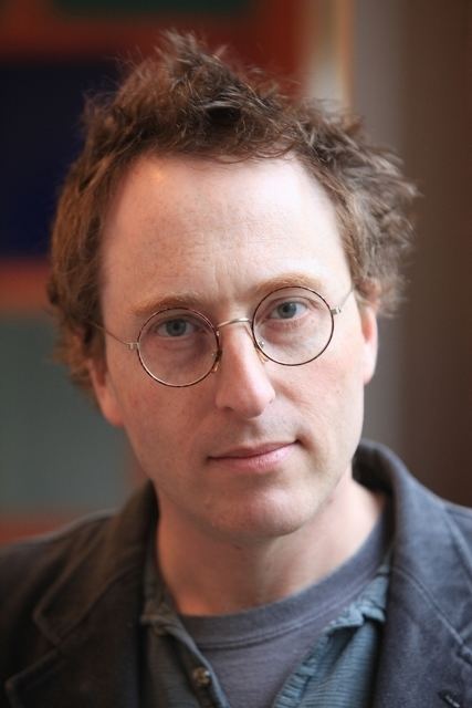 Jon Ronson is serious, has brown hair, wears black eyeglasses, and a gray and green shirt under a black suit.