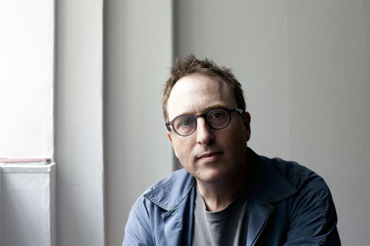 Jon Ronson is serious, has brown hair, and wears black eyeglasses, and a gray shirt under a blue jacket.
