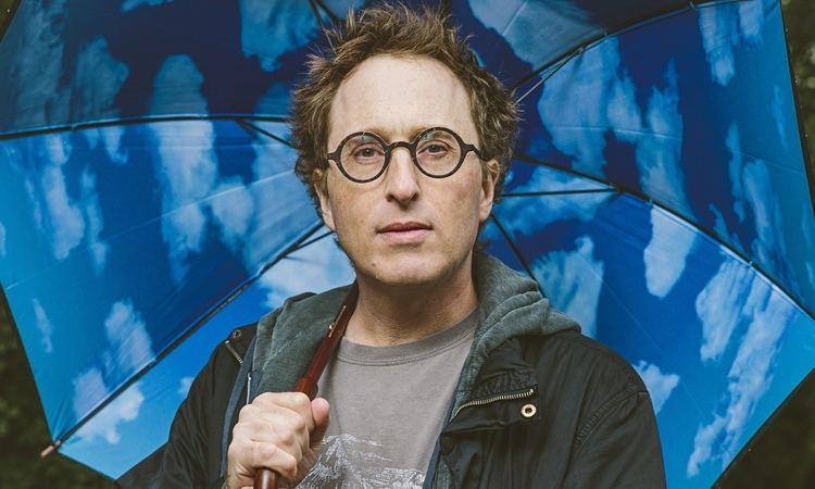 Jon Ronson is serious, has brown hair, his right hand holds a blue umbrella with a sky design, and wears a gray shirt with a print under a black jacket.