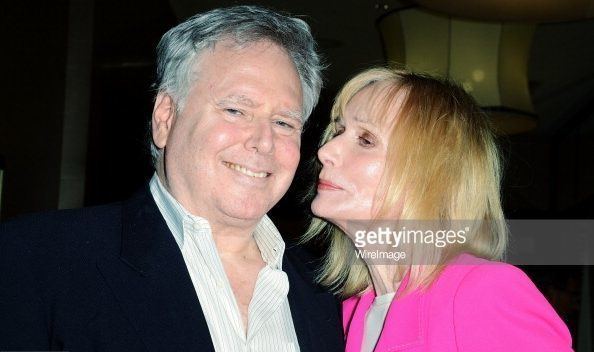 Jonathan D. Krane smiling while Sally Kellerman is going to kiss his cheek. Jonathan is wearing a black coat over white striped long sleeves while Sally with blonde hair and wearing a pink coat over a white top.