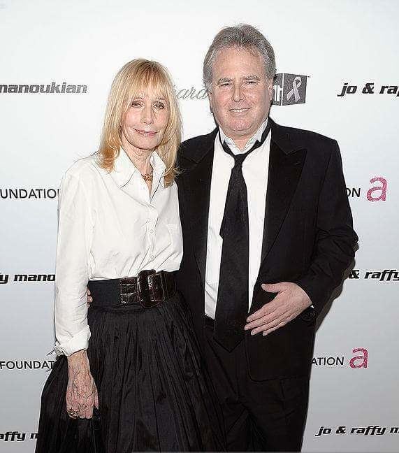 Jonathan D. Krane is smiling while Sally Kellerman with a tight-lipped smile. Jonathan is wearing a black coat over white long sleeves, a black necktie, and black pants while Sally with blonde hair, wearing a necklace, a ring, a white long sleeve top, a black belt, and a black skirt.