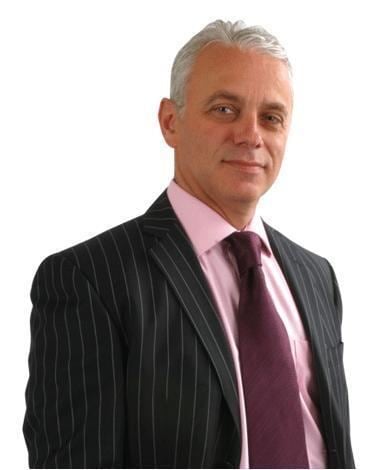 Jonathan Gilbert with a tight-lipped smile while wearing a pink long sleeve under a violet necktie and black striped coat