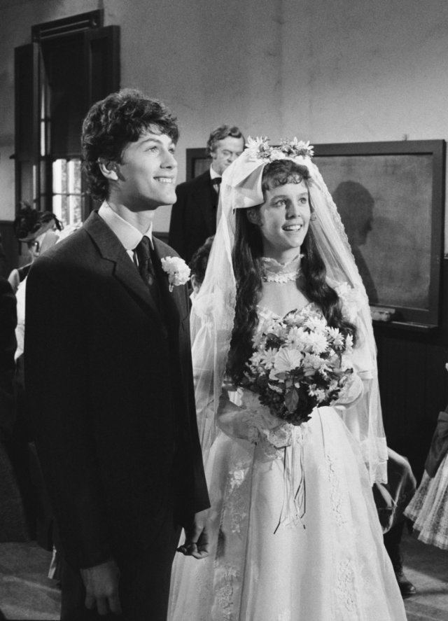 Jonathan Gilbert and Sherri Stoner are smiling while looking at something and Sherri holding a bouquet of flowers. Jonathan wearing pants and long sleeves under a necktie and coat while Sherri wearing long sleeve wedding down and veil in a scene from the 1974 TV Series, Little House on the Prairie