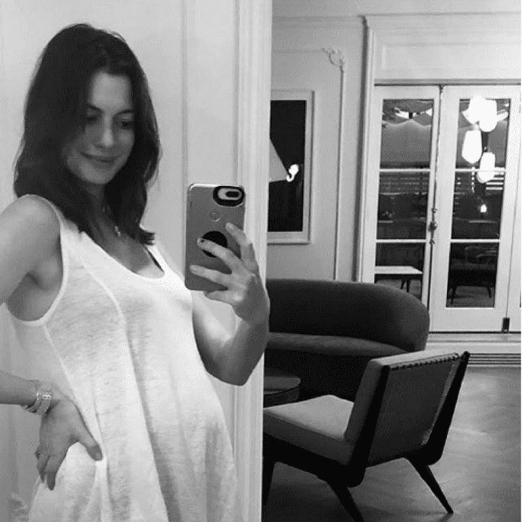 Anne Hathaway smiling and taking a mirror selfie while showing her baby bump and she is wearing a sleeveless dress