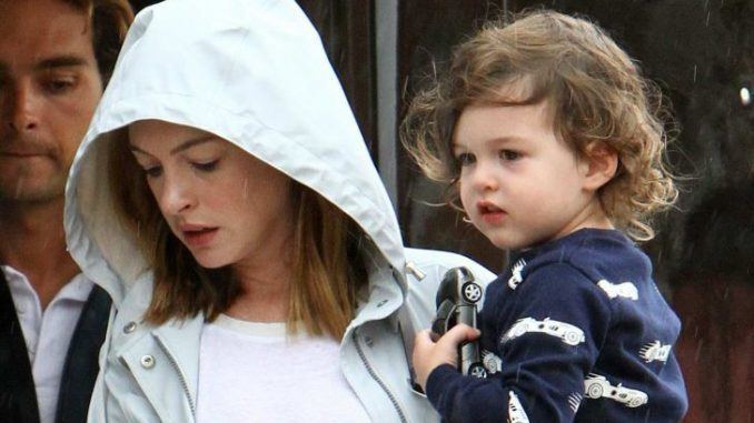 Anne Hathaway carrying her son Jonathan Rosebanks Shulman while Anne is wearing a gray jacket and white blouse and her son is wearing a blue sweatshirt