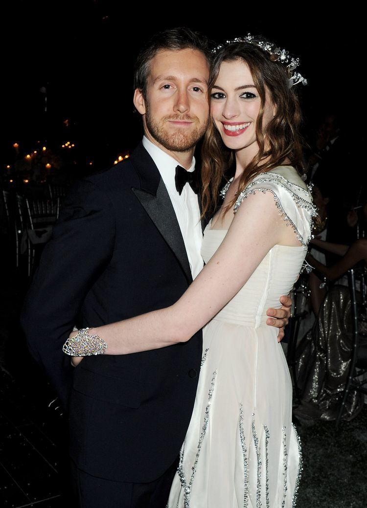 Adam Shulman and Anne Hathaway are smiling and hugging each other while Adam wearing a black coat and long sleeves and Anne wearing a white dress