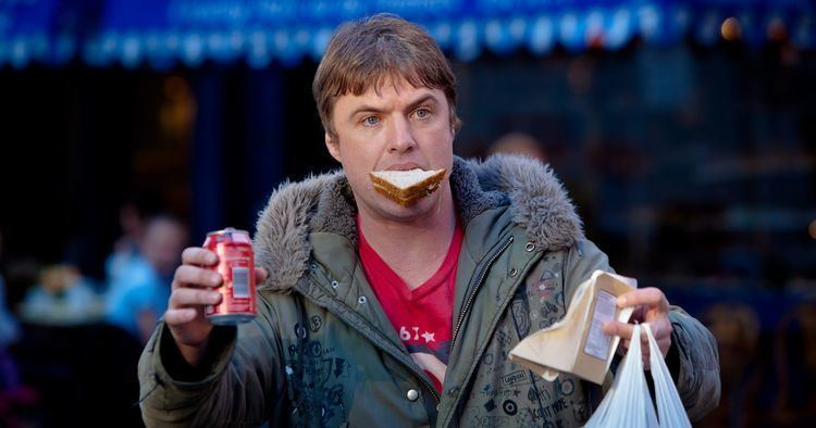 Jonny Owen wearing red shirt and jacket with sandwich on his mouth and holding a drink in can and plastic bag