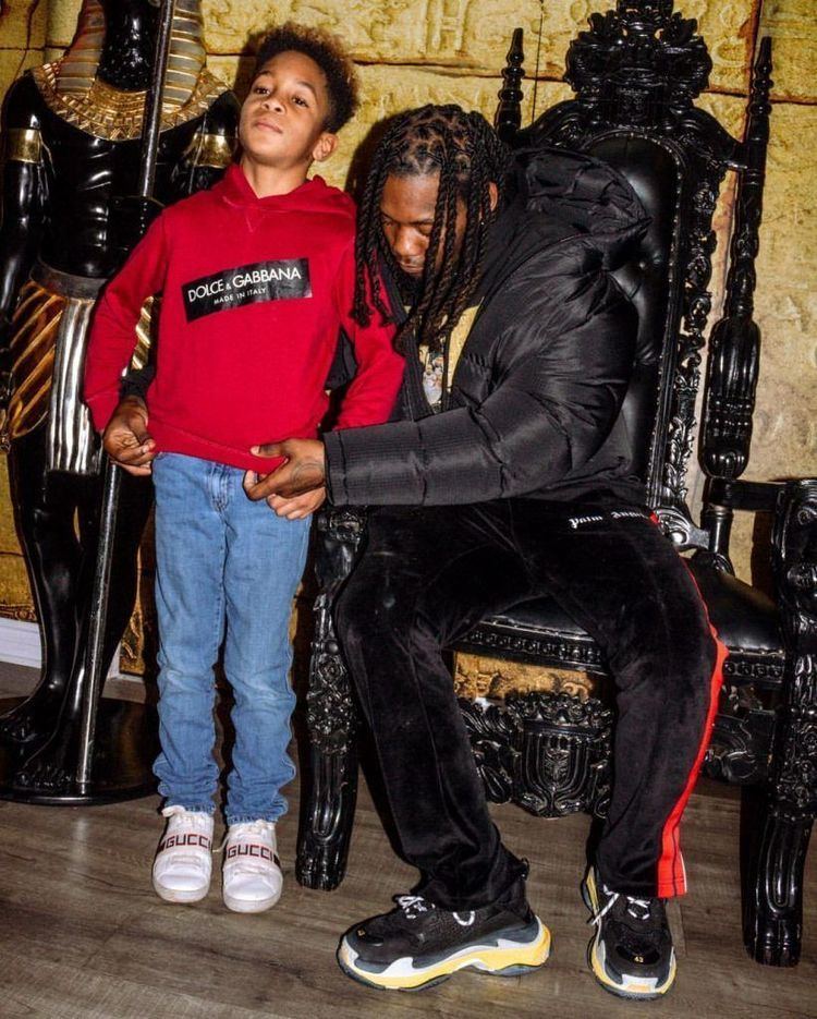 Jordan Cephus standing and wearing a red hoodie jacket, blue jeans, and white shoes while his father, Kiari helps him fix his jacket