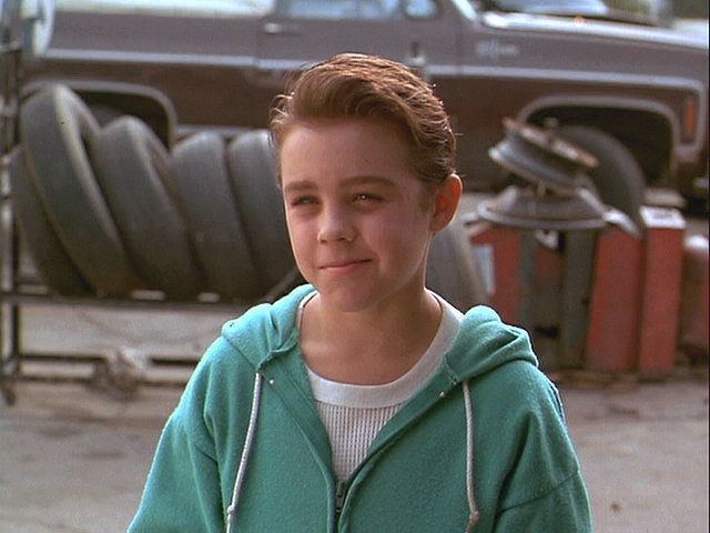 Jordan Christopher smirking wearing a white shirt and a green jacket in a movie scene from "Motorama (1991)"