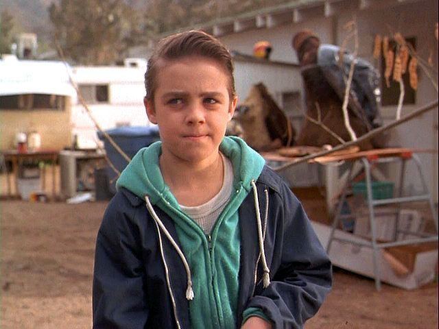 Jordan Christopher wearing a white shirt and a green jacket outdoor in a movie scene from "Motorama (1991)"