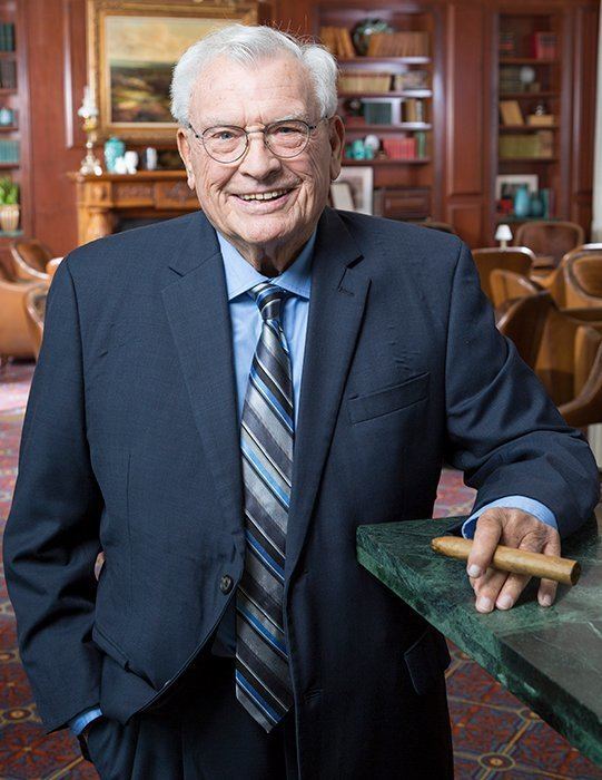 Joseph A. Hardy III wearing eyeglasses, black coat, blue long sleeves, blue and gray neck tie while holding tobacco