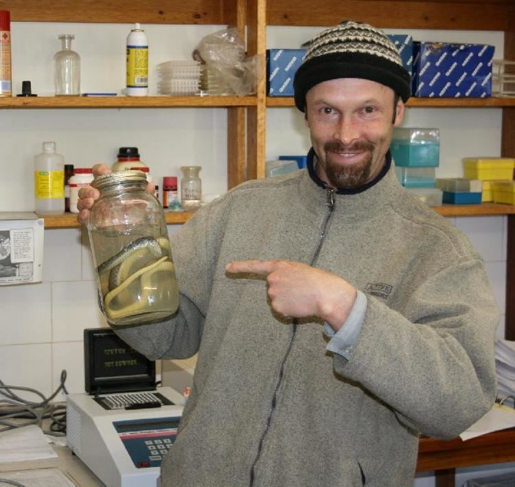 A man smiling while pointing at the jar with a snake inside and wearing a gray jacket and beanie